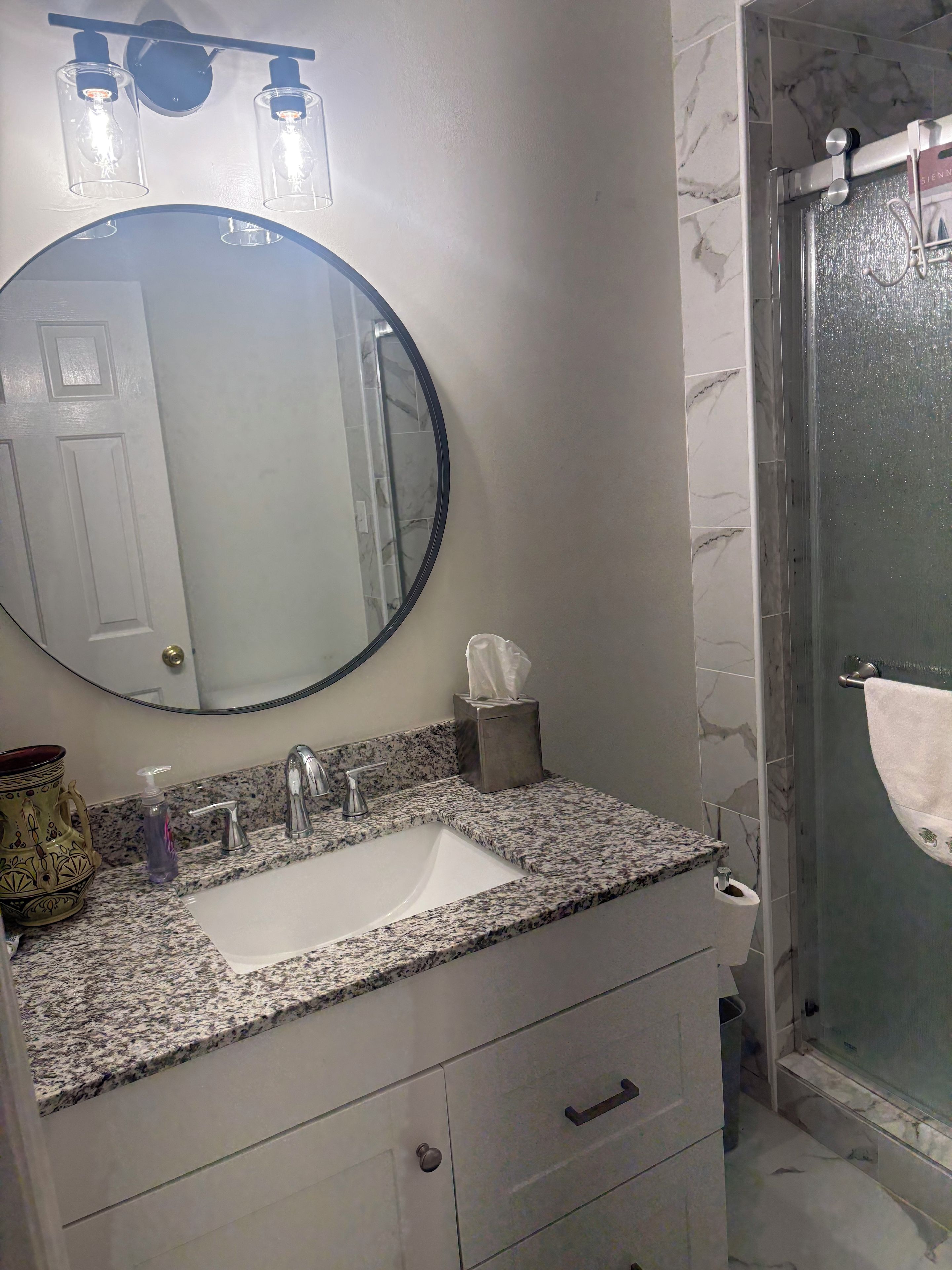A bathroom with a marble-tiled shower, a sink with a marble countertop, a round mirror, and a towel rack.