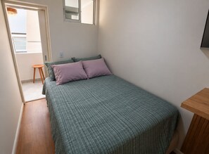 Ein Einzelbett mit einer grün-weißen Karomatik, zwei Kissen, einem Nachttisch aus Holz und einem kleinen Hocker.