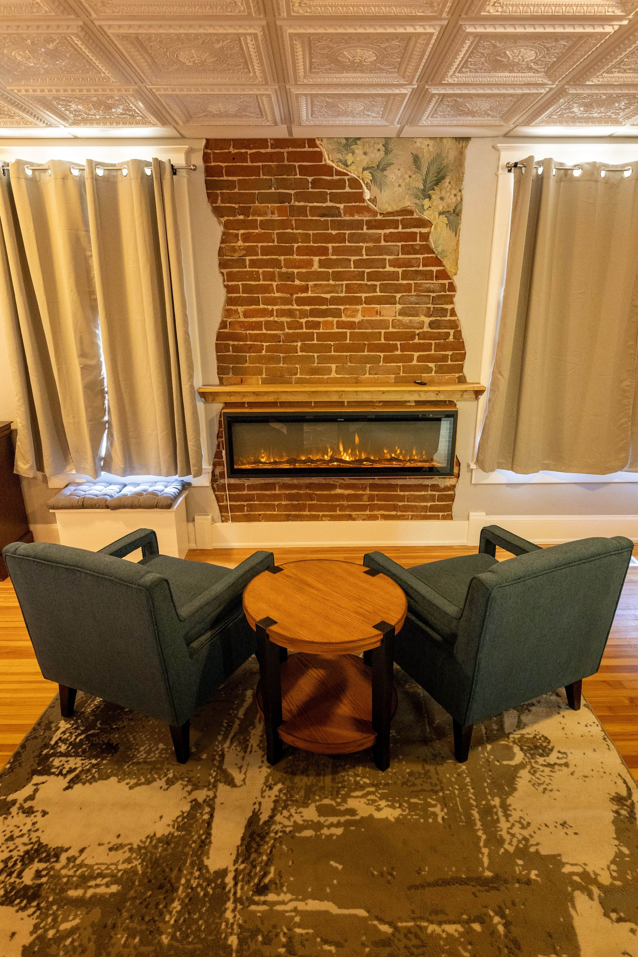 A room with a brick fireplace, two armchairs, a round wooden table, and a patterned rug.