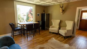 A room with a wooden floor, a dining table with chairs, a white armchair, a large window with a view of trees, and a mounted animal head on the wall.
