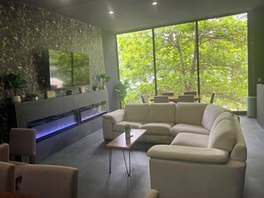 Una sala de estar moderna con un sofá modular, un televisor de pantalla plana montado en la pared, una chimenea empotrada y un amplio ventanal con vistas a exuberante vegetación.