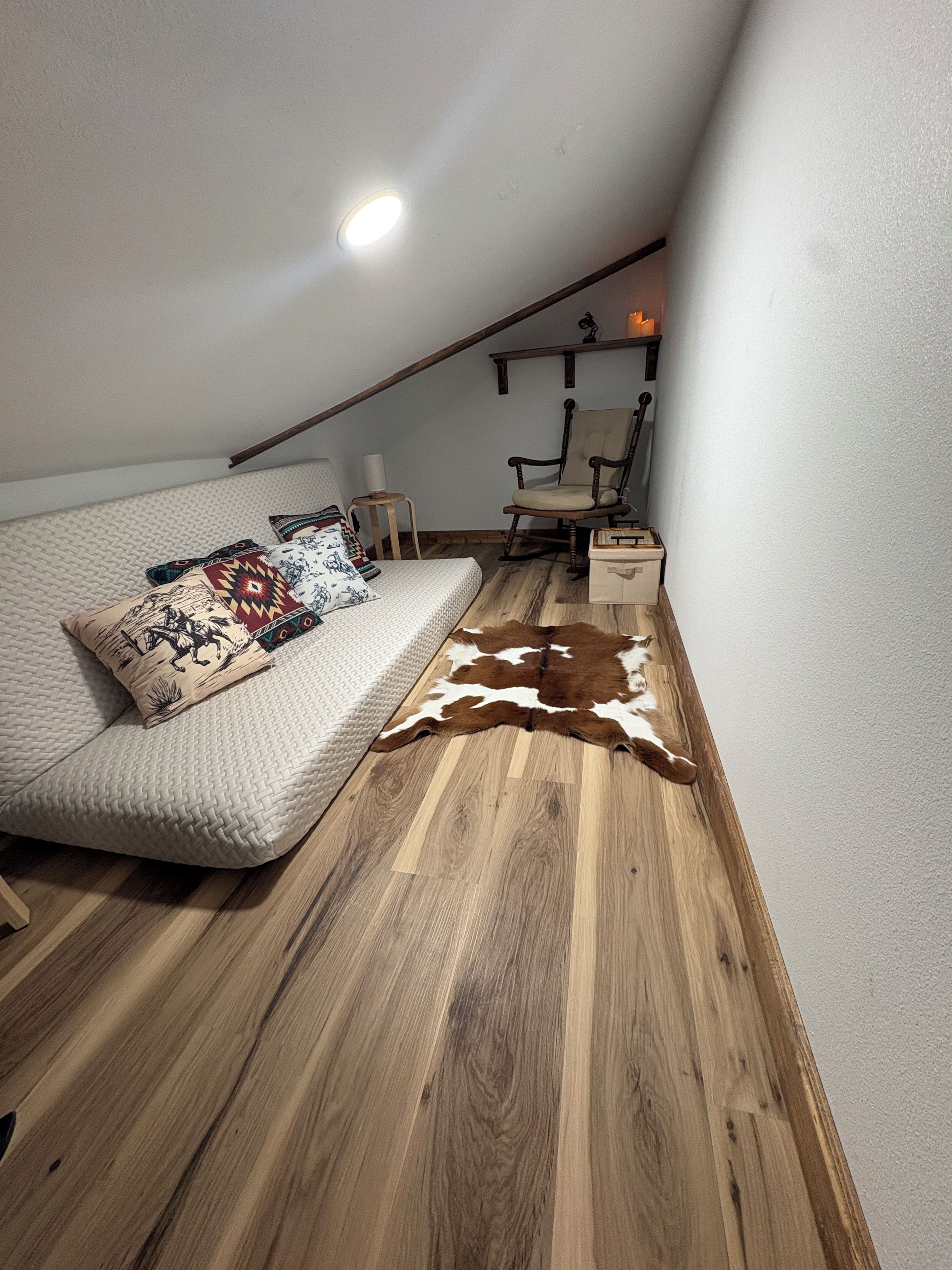 Une chambre mansardée chaleureuse avec un plafond en pente, un canapé-lit agrémenté de coussins à motifs, un plancher en bois, un tapis en peau de vache, une chaise et une petite table.