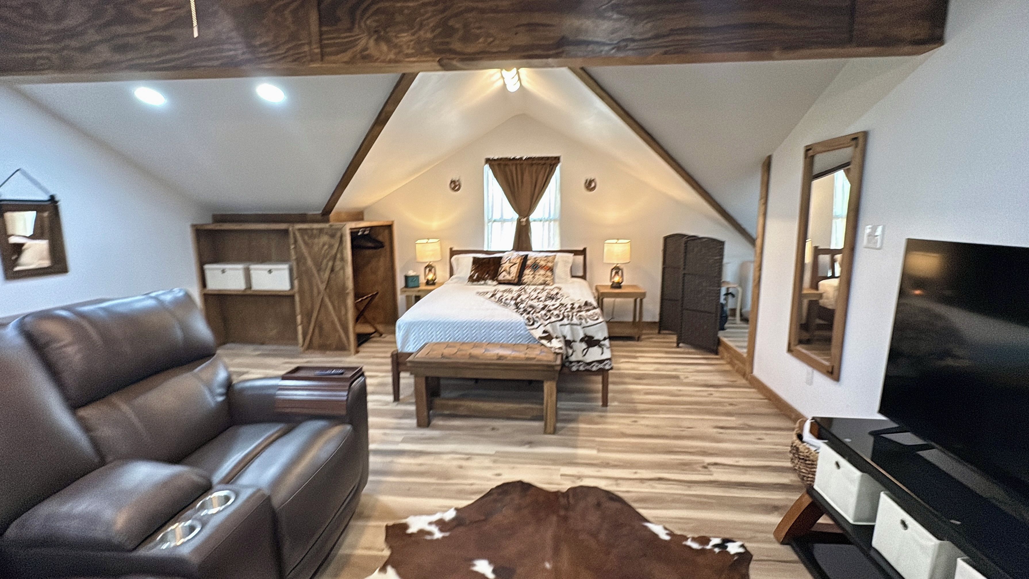 A loft bedroom with a sloped ceiling, a bed with a patterned bedspread, a leather sofa, a wooden bench, and a television on a stand.