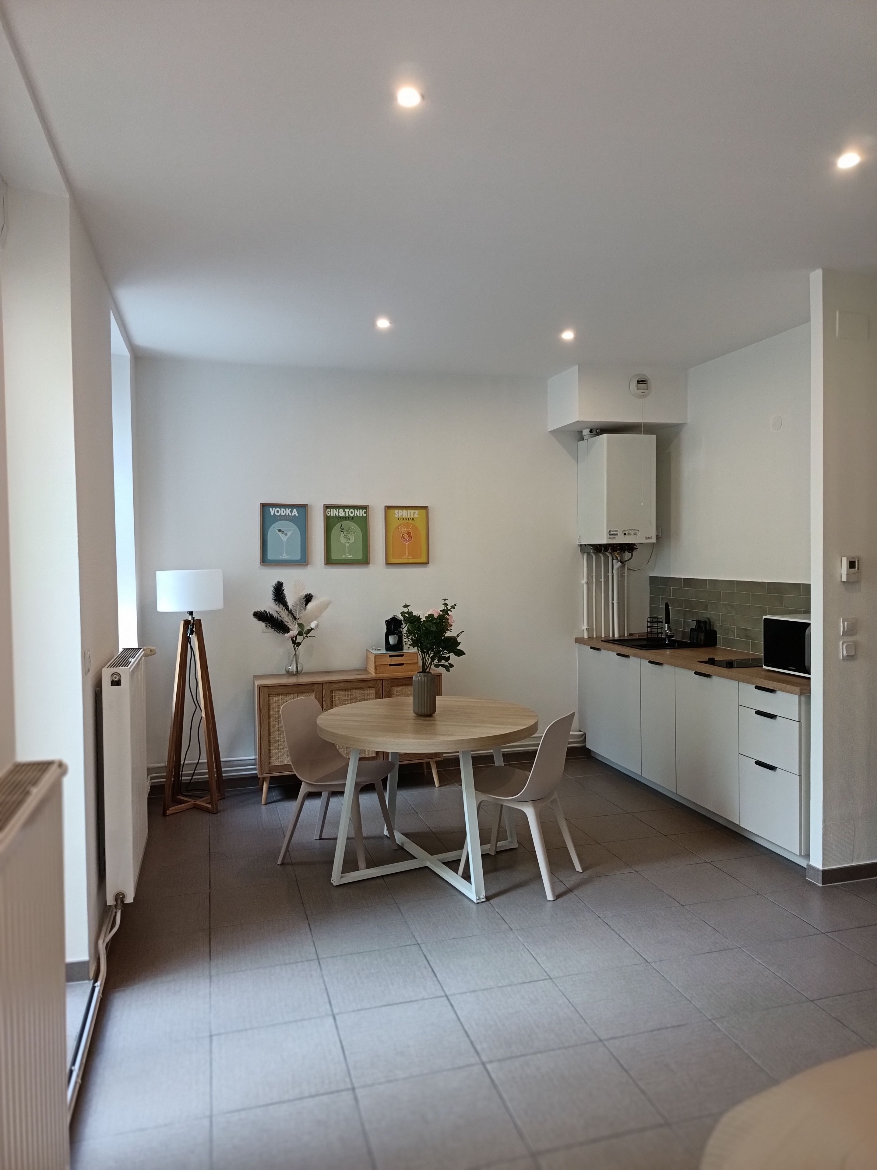 Une cuisine moderne avec des meubles blancs, une table à manger avec des chaises et un petit îlot central agrémenté d’une plante. Des tableaux encadrés ornent le mur et un radiateur est posé sur le sol.