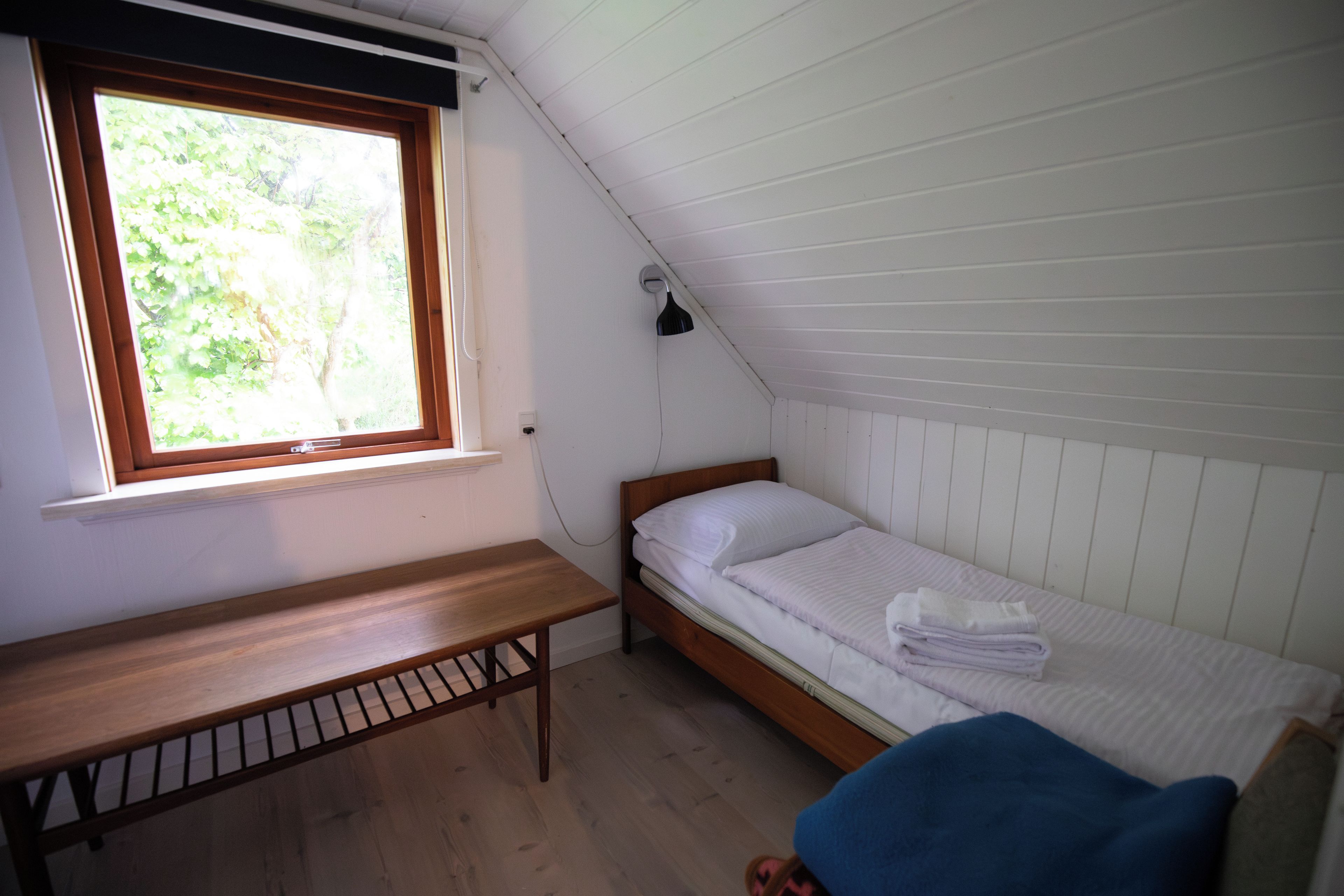 Et lille værelse med en enkelt seng, en træbænk, et vindue med udsigt til grøn vegetation og en lampe på væggen.