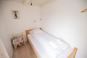 Una habitación pequeña con cama individual, estructura de madera, colchón blanco y mesita de noche con lámpara.