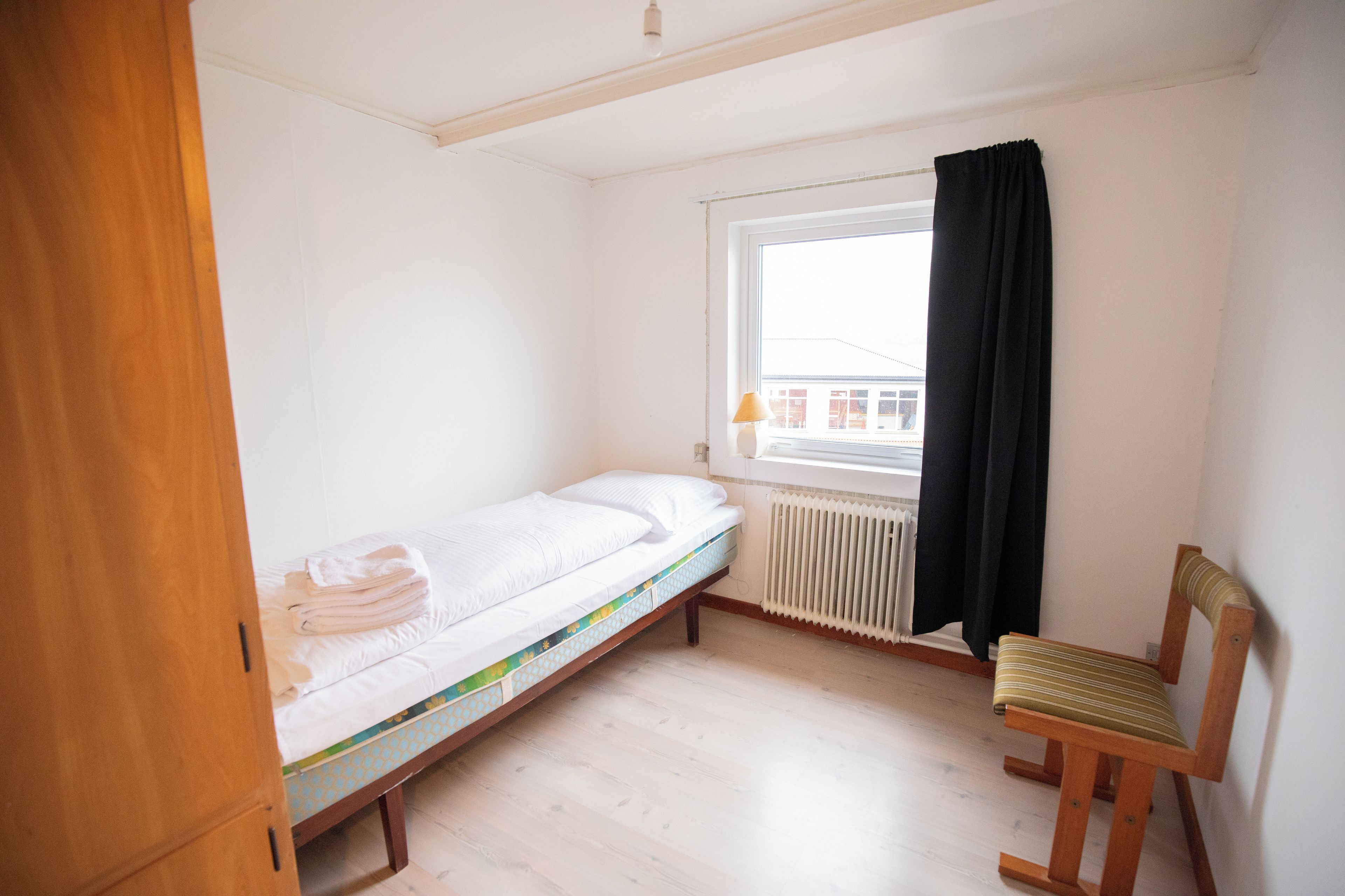 A small, single-bed room with a wooden floor, a radiator, a window with a black curtain, and a wooden chair.