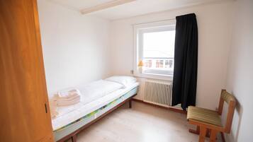 A small, single-bed room with a wooden floor, a radiator, a window with a black curtain, and a wooden chair.