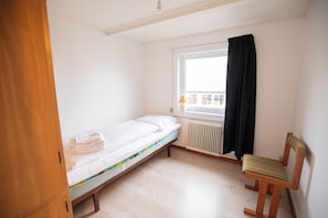 A small, single-bed room with a wooden floor, a radiator, a window with a black curtain, and a wooden chair.