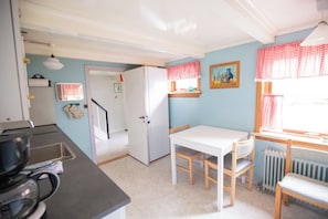 Cocina compacta con refrigerador, lavamanos y estufa. Hay una mesa de comedor con sillas y una ventana con cortinas a cuadros.