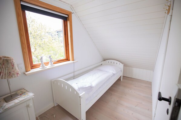 A small, single-bed room with a window, a bedside table, and a door.