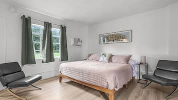 A bedroom with a bed, two chairs, a window with curtains, a wall-mounted air conditioner, and a framed picture.
