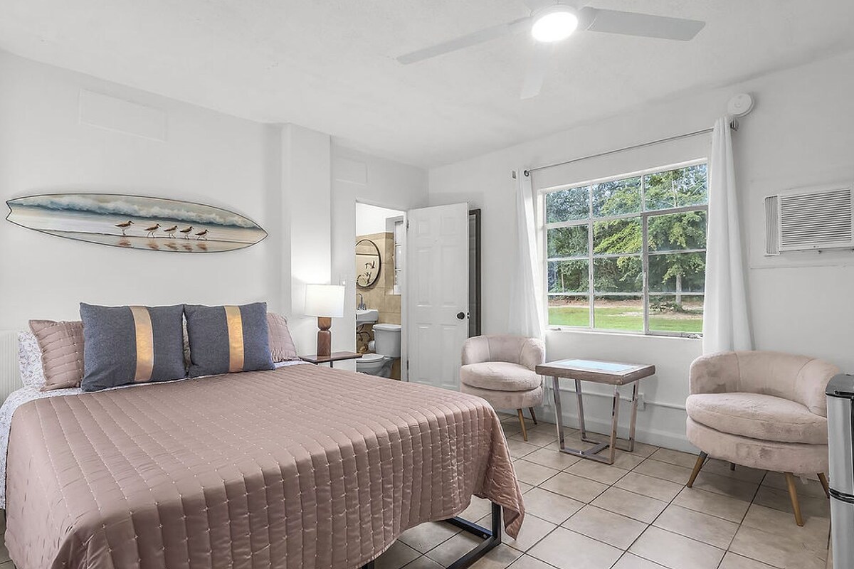 A bedroom with a bed, a chair, a table, a window, and a ceiling fan.