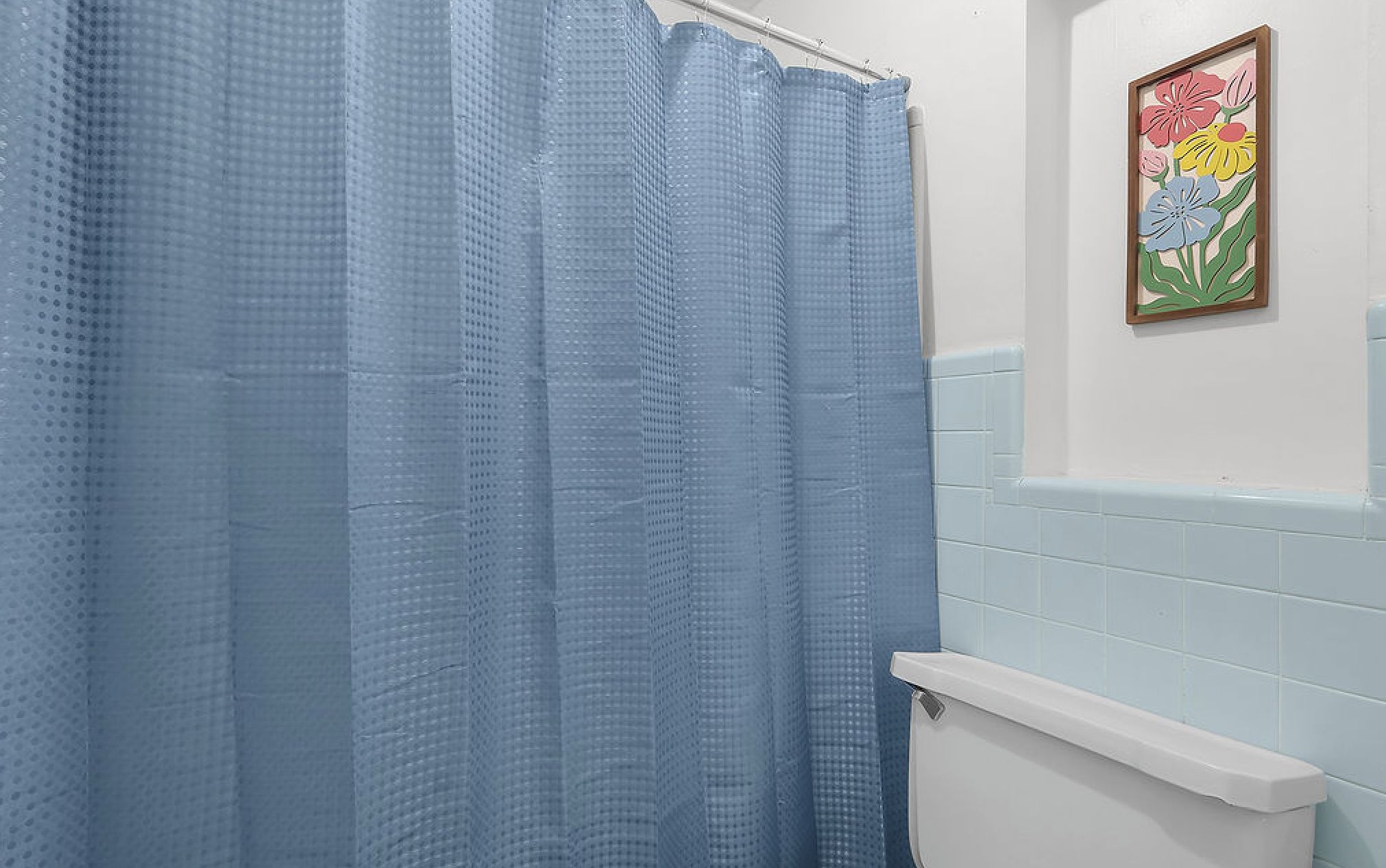 Um banheiro com cortina de chuveiro azul, vaso sanitário branco e um quadro de flores na parede.