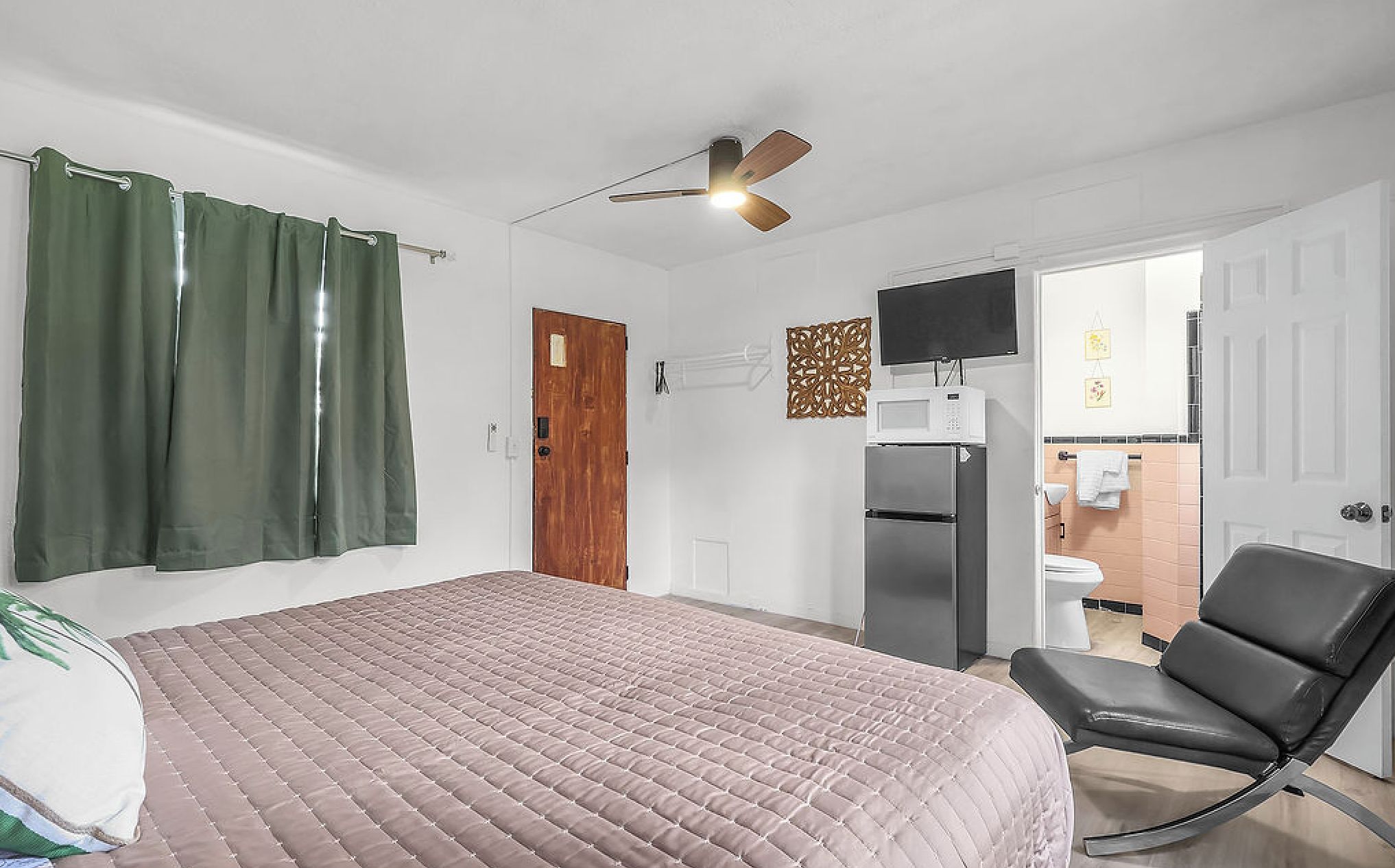 A bedroom with a bed, a chair, a ceiling fan, a door, and a bathroom visible through an open door.