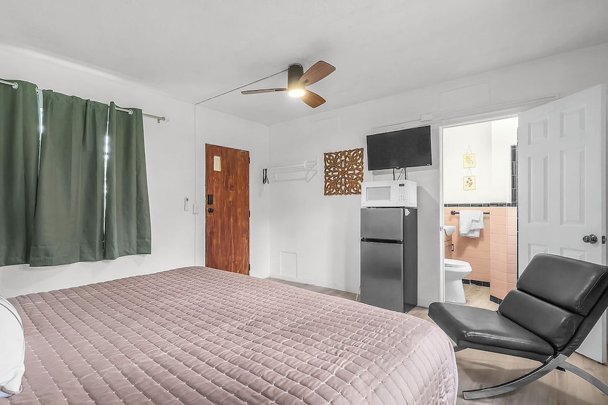 A bedroom with a bed, a chair, a ceiling fan, a door, and a bathroom visible through an open door.