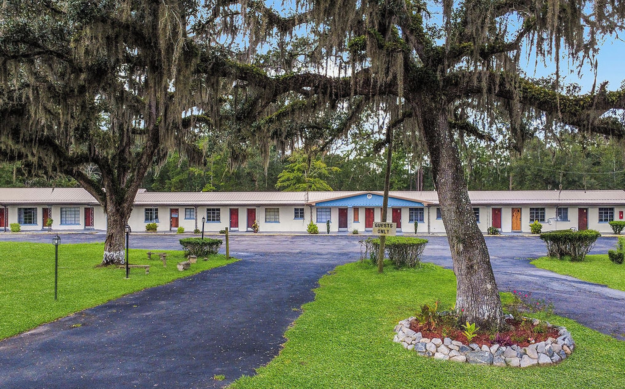 A motel with a central driveway, surrounded by well-maintained lawns and trees with hanging moss.
