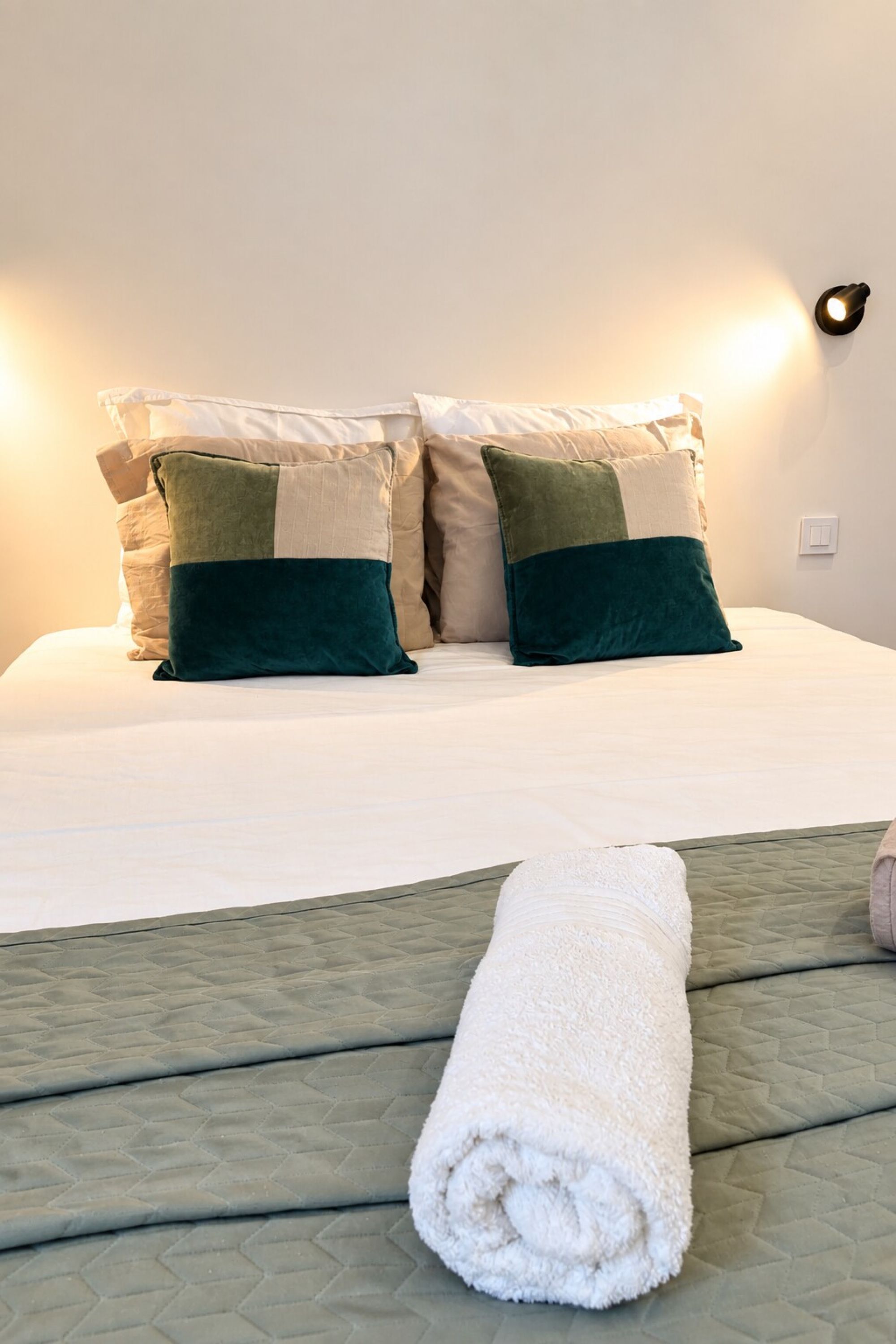 A neatly made bed with a white duvet, two pillows with a green and beige color scheme, and a rolled white towel on the bed.