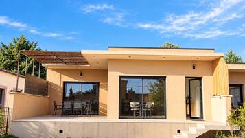 A modern two-story house with a flat roof, large sliding glass doors, and a patio area with outdoor furniture.