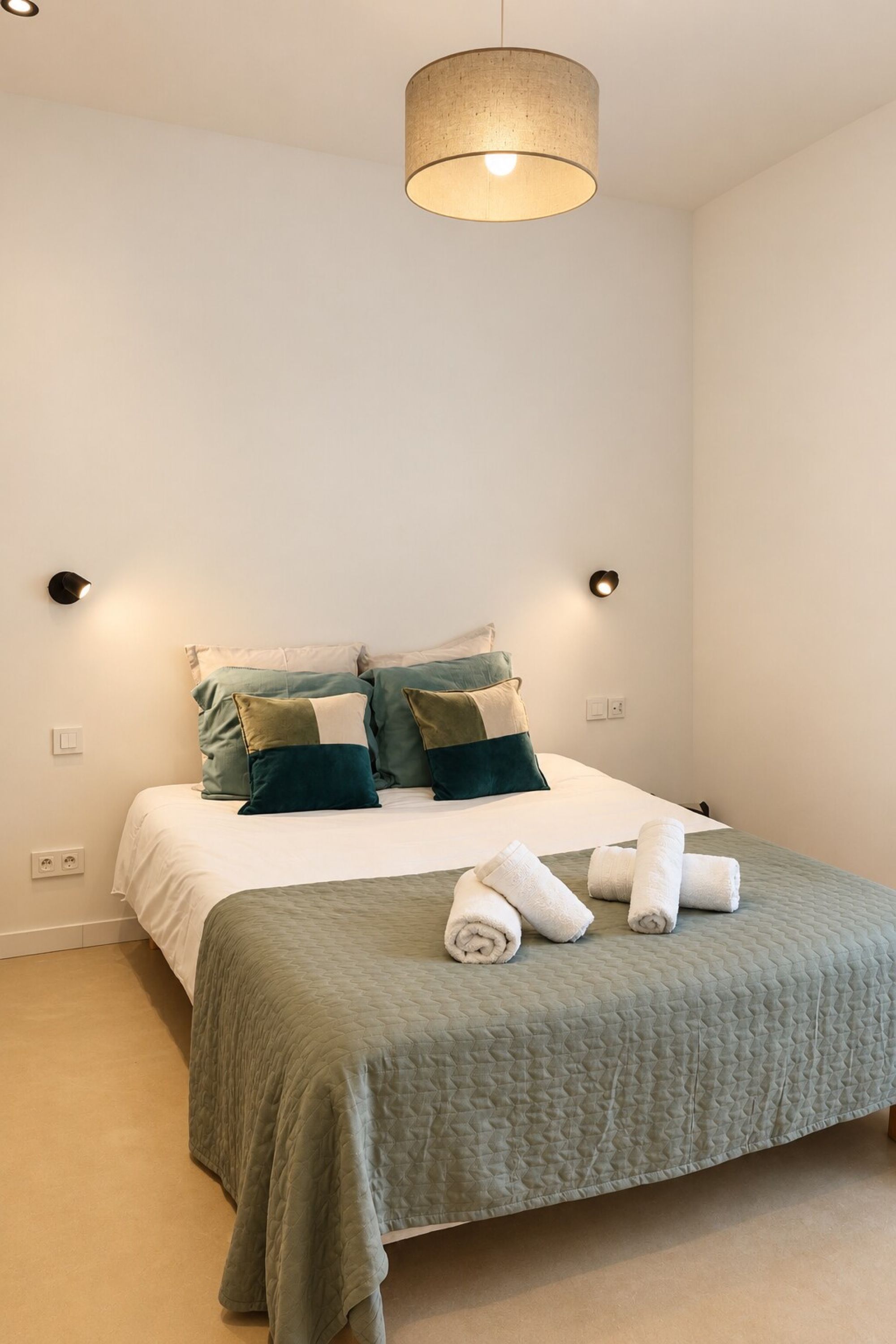 A neatly made bed with a green bedspread, two pillows, and rolled towels.