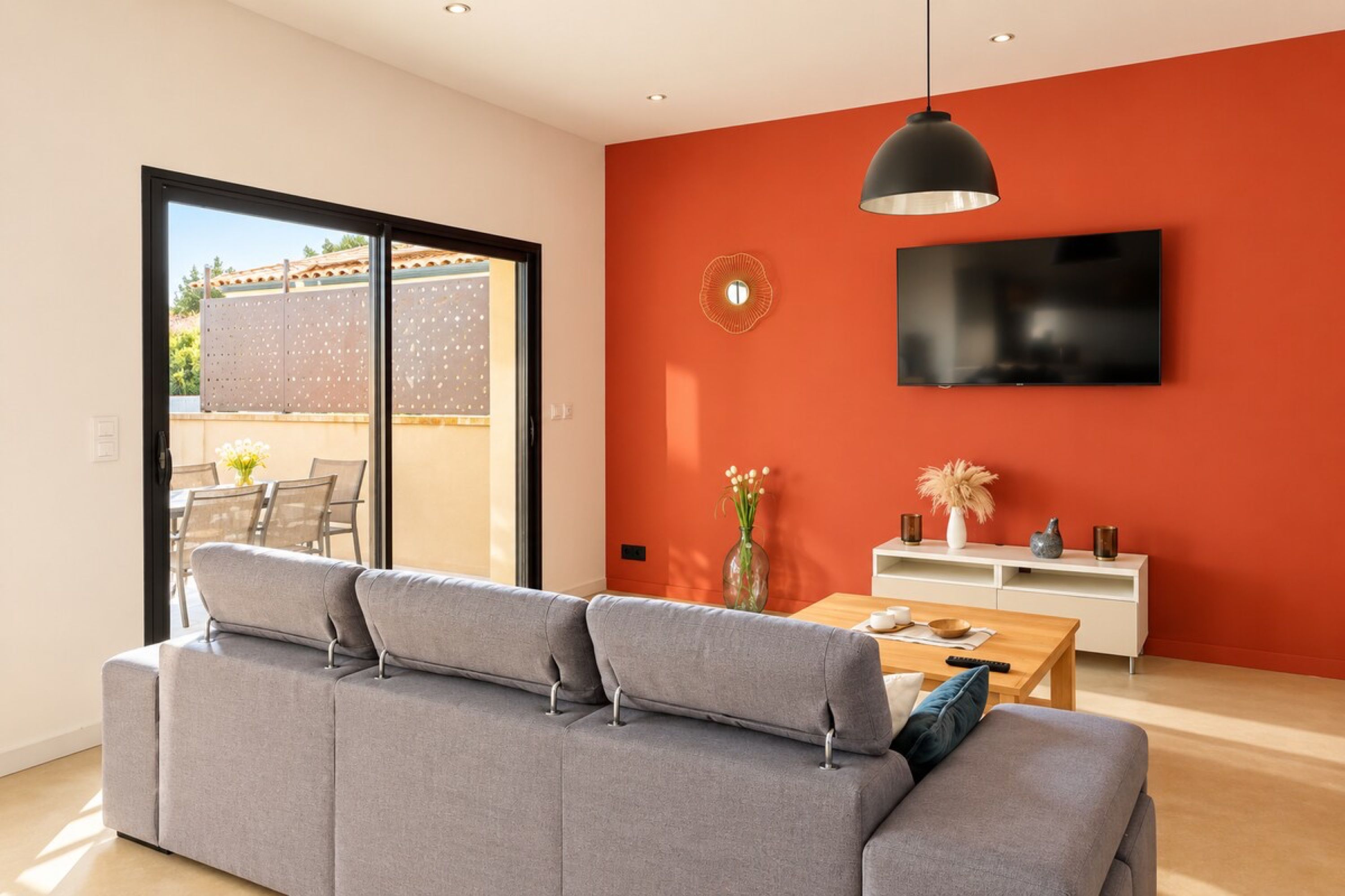 Una sala moderna con un sofá gris, una mesa de centro de madera, un televisor de pantalla plana montado en una pared roja y un ventanal corredero que da a un área exterior.