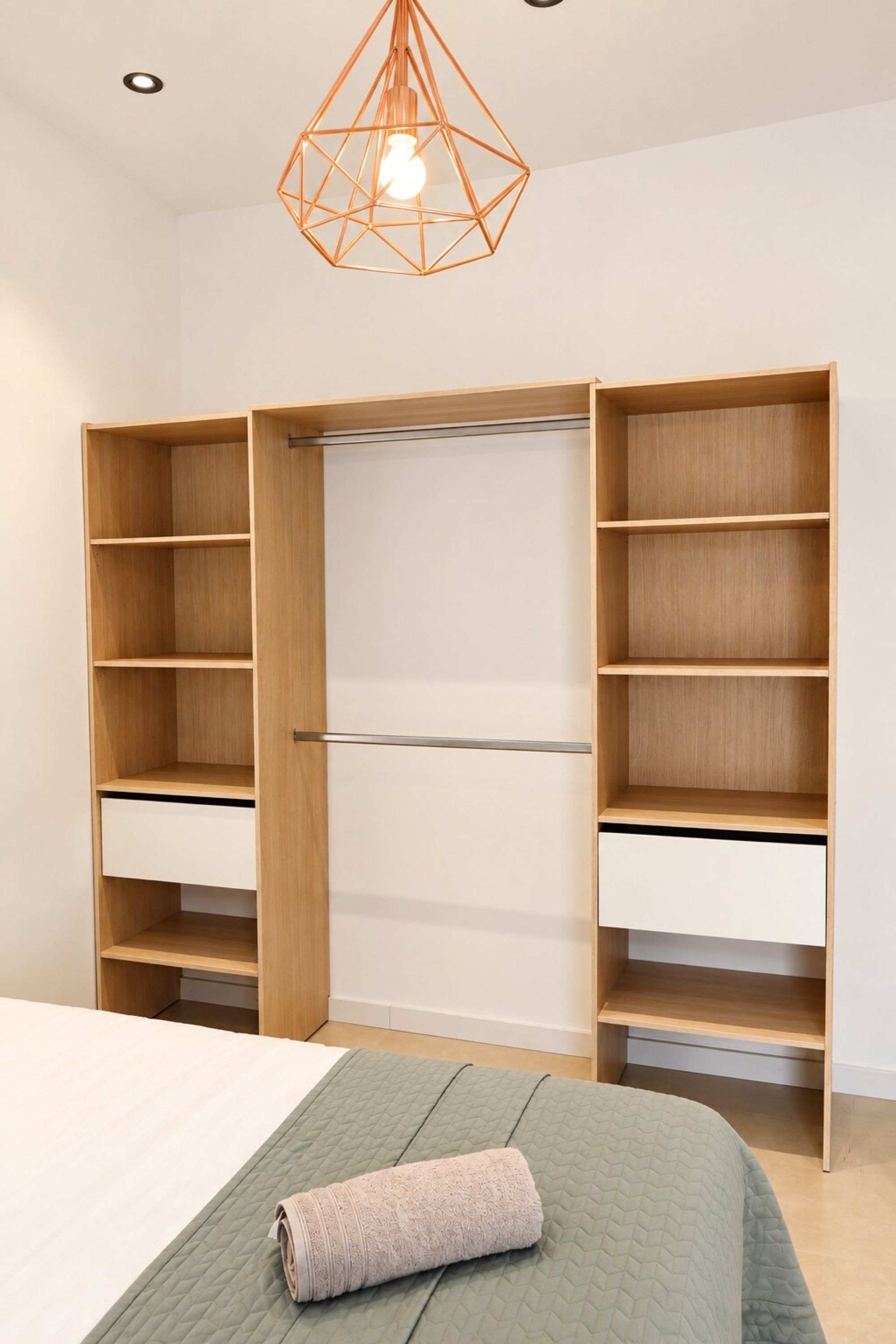 Ein Schlafzimmer mit einem Bett, einer grünen gesteppten Bettdecke, einem aufgerollten Handtuch, einem hölzernen Kleiderschrank mit mehreren Einlegeböden und einer geometrischen Pendelleuchte.