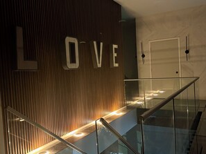 Un intérieur moderne avec un escalier en verre, un mur en bois sur lequel le mot « AMOUR » est illuminé, et une porte sur la droite.