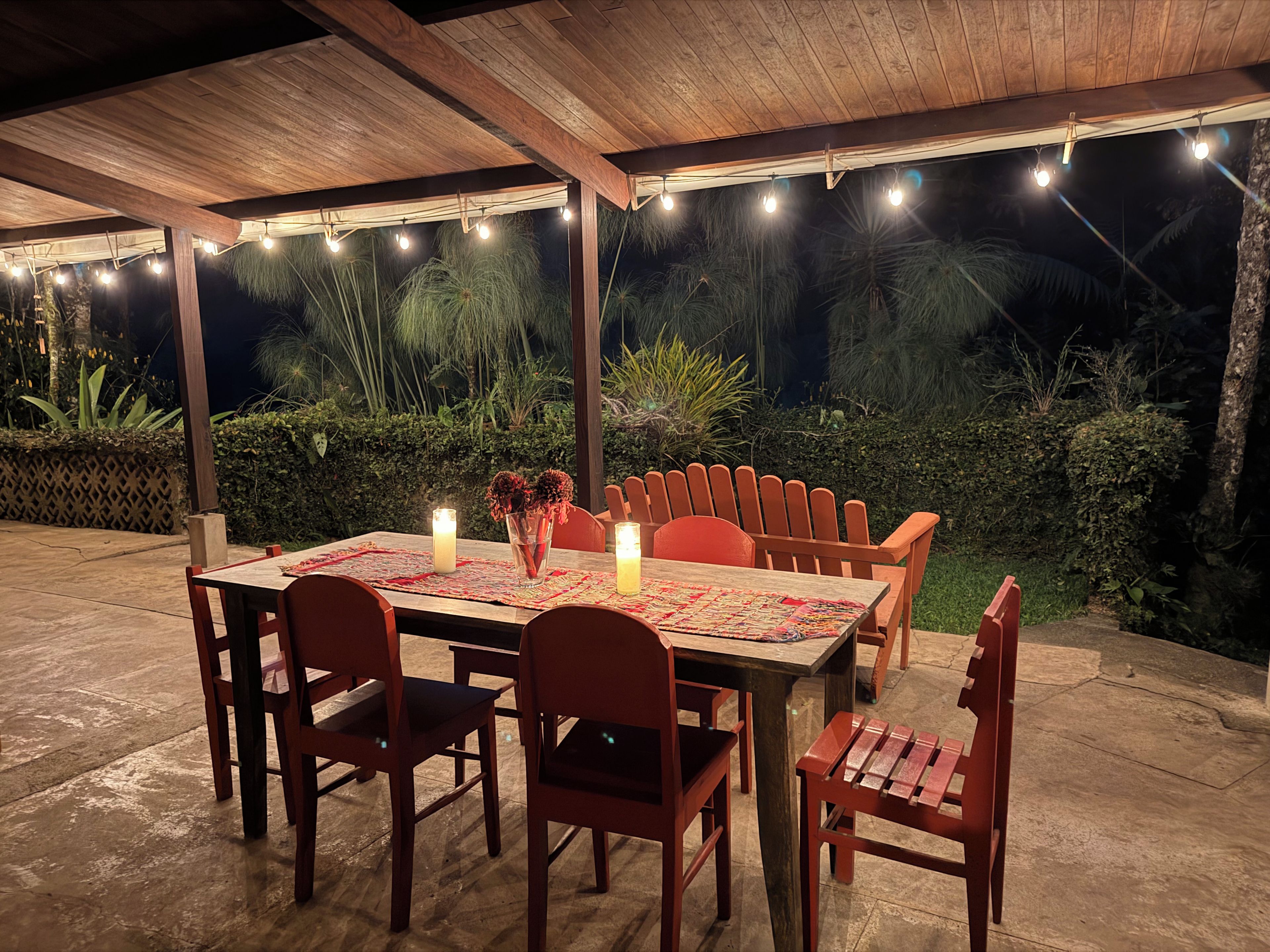 An outdoor dining area with a wooden table and chairs, string lights, and a view of greenery.