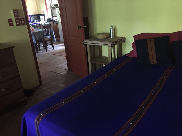 A room with a bed, a table with a metal container, and a chair. There is a door leading to a kitchen area.