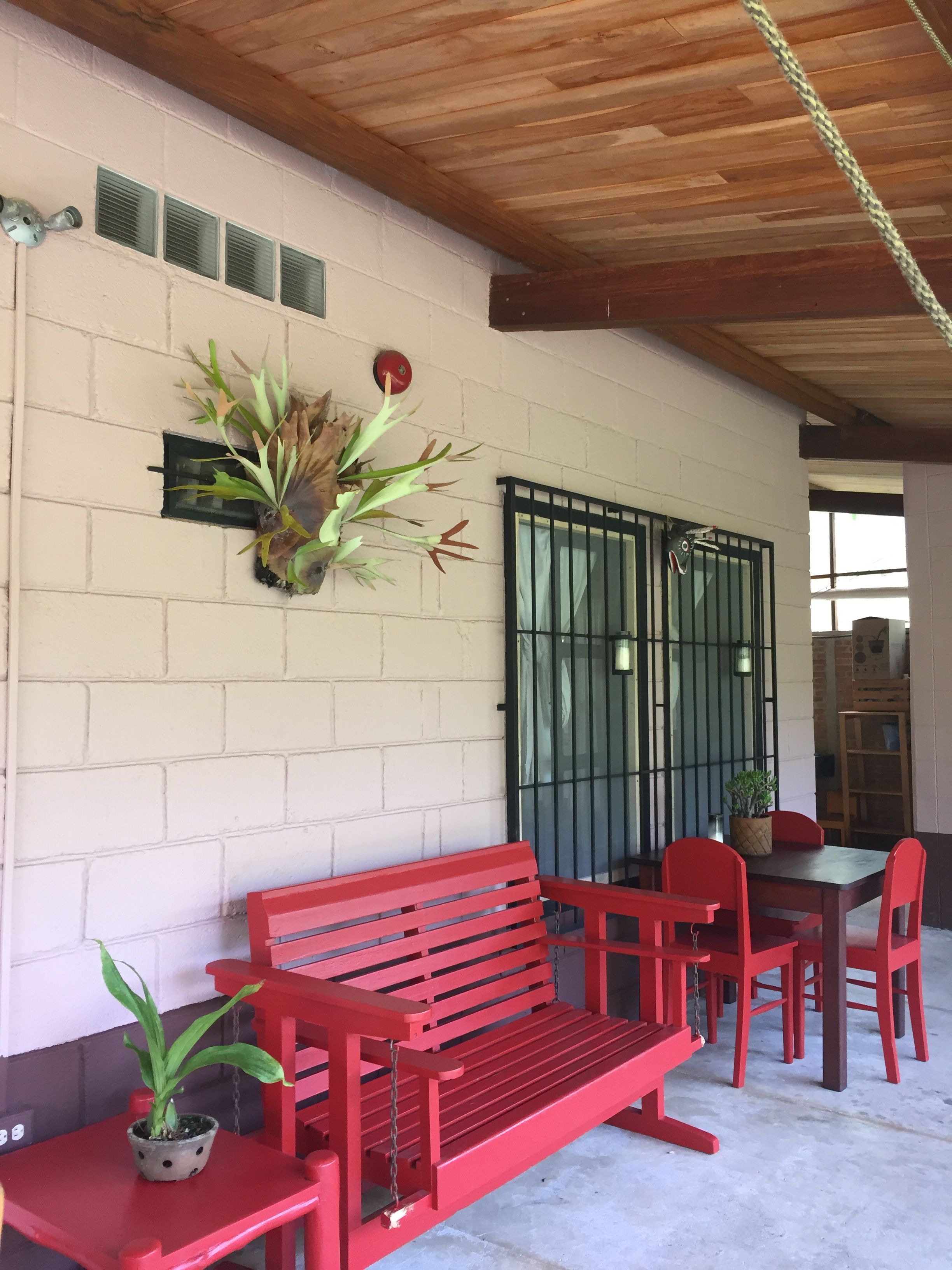 A red bench and chairs in a covered outdoor area with a brick wall, a metal gate, and a potted plant.