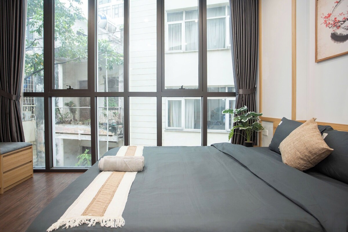 Een slaapkamer met een groot raam, een bed met een grijze matras en kussens, een houten nachtkastje en een pot met een plant.
