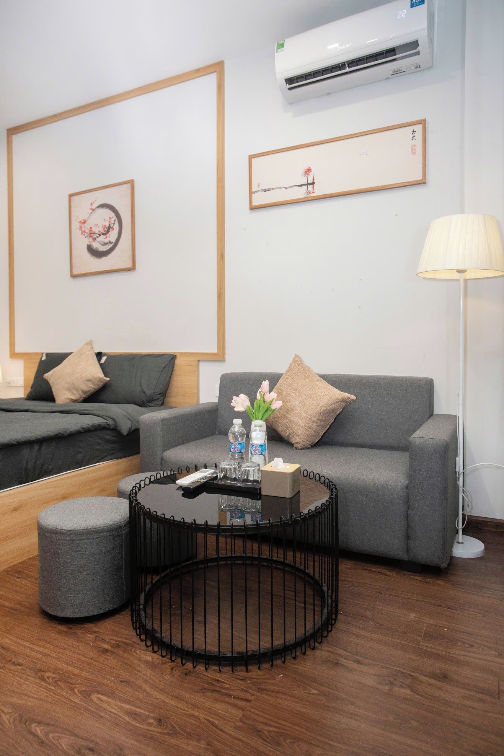 A modern hotel room with a grey sofa, a round black coffee table with amenities, a grey ottoman, and a floor lamp.