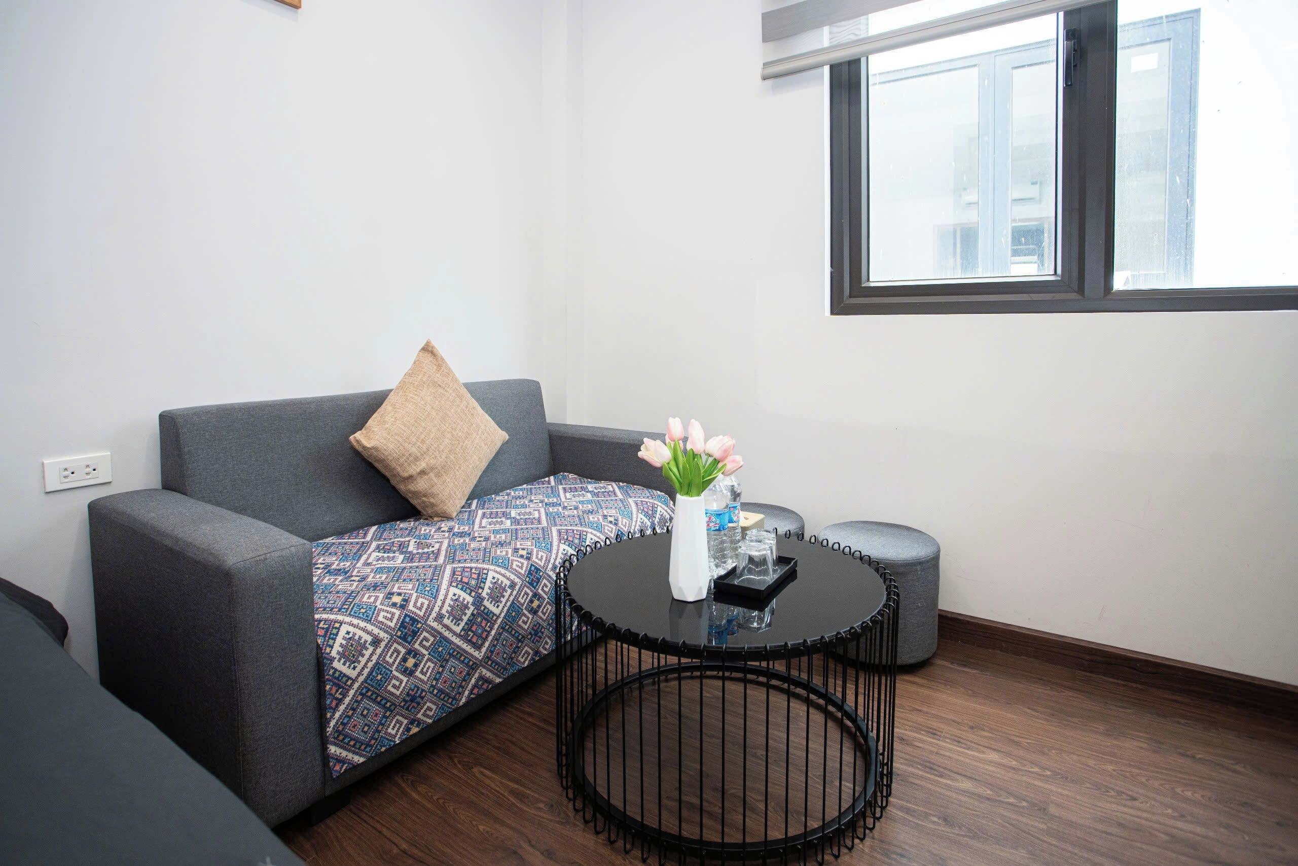 A modern living room with a grey sofa, patterned throw, a small round table with a vase of flowers, and a window with blinds.