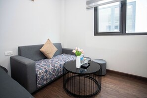 Ein modernes Wohnzimmer mit einem grauen Sofa, einem gemusterten Überwurf, einem kleinen runden Tisch mit Blumenvase und einem Fenster mit Jalousien.