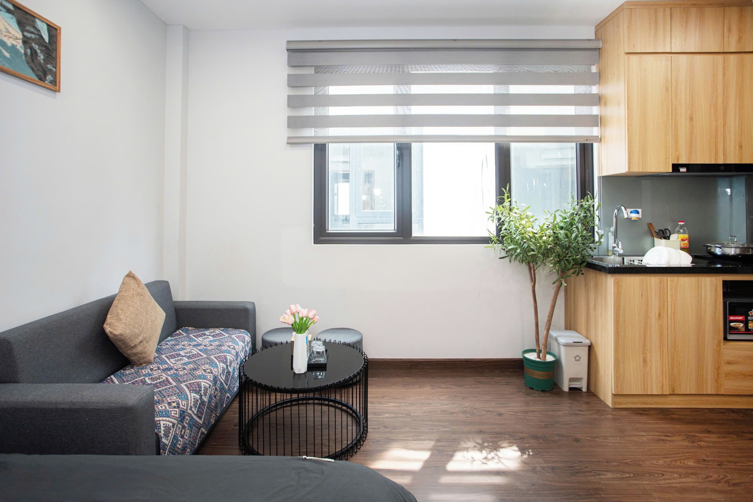 Un espace de vie compact comprenant un canapé, une petite table ronde agrémentée d’un vase de fleurs, un coin cuisine avec une cuisinière et des placards, et une fenêtre avec des stores.