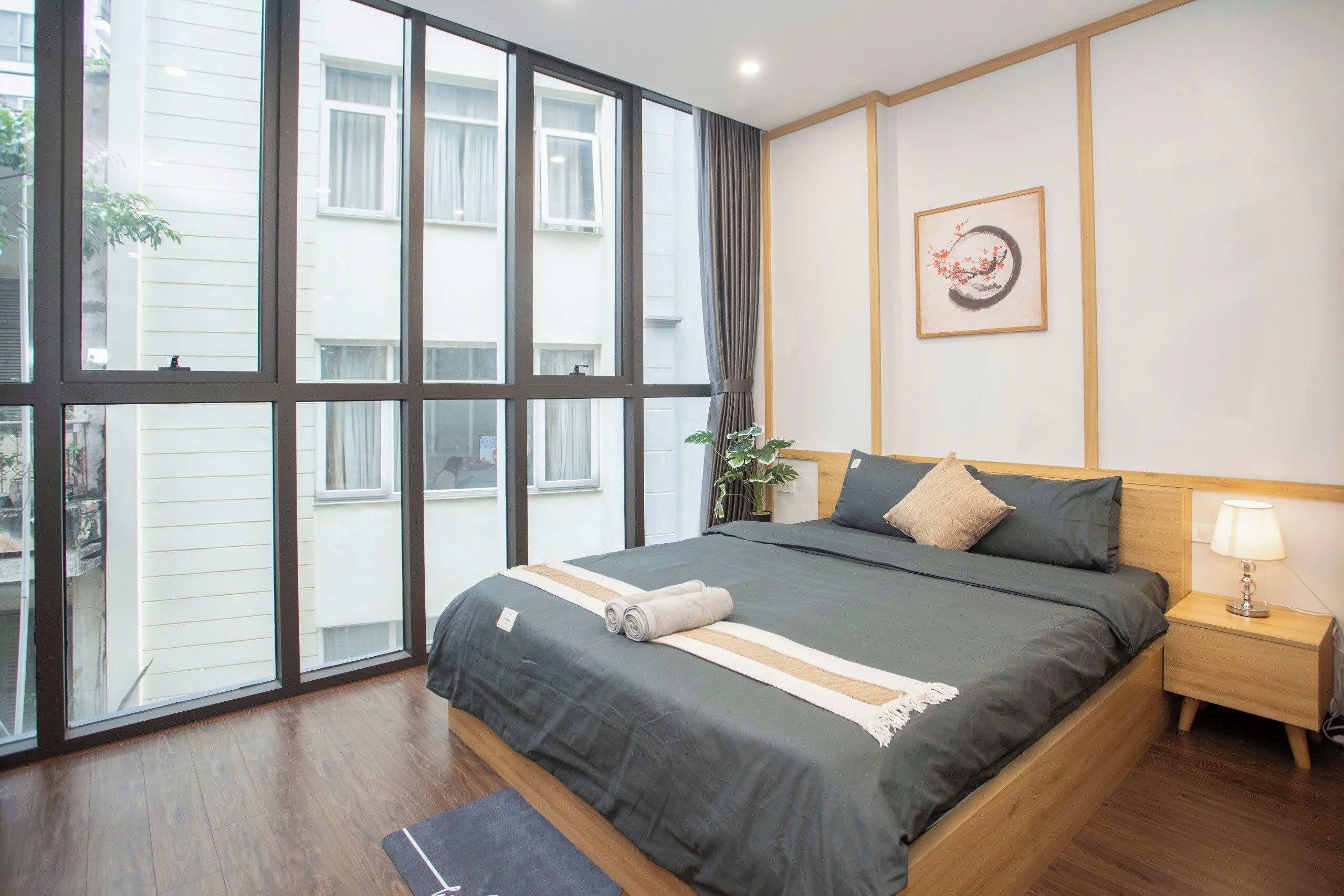 A bedroom with a large window, a bed with grey bedding, a bedside table with a lamp, and a wall-mounted artwork.