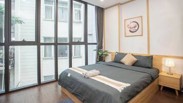 A bedroom with a large window, a bed with grey bedding, a bedside table with a lamp, and a wall-mounted artwork.