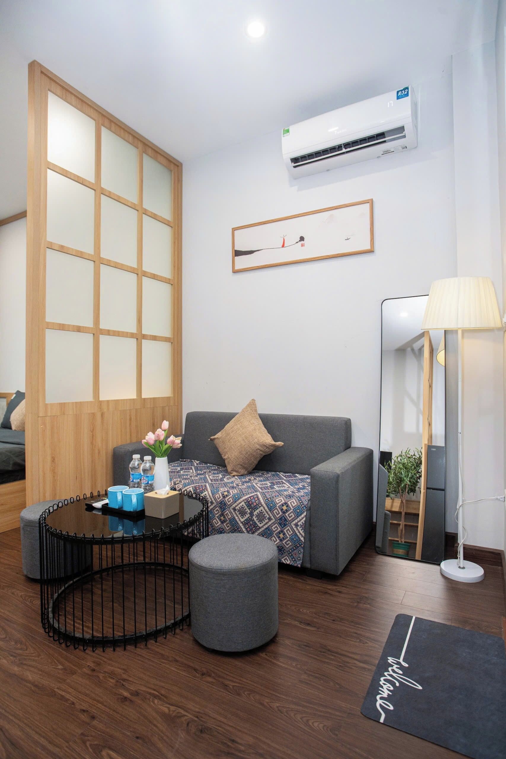 A modern living room with a grey sofa, a round coffee table, a small ottoman, a wooden partition, an air conditioning unit, a framed artwork, and a floor lamp.