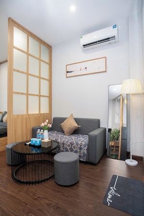 Ein modernes Wohnzimmer mit einem grauen Sofa, einem runden Couchtisch, einem kleinen Hocker, einer Holzabtrennung, einer Klimaanlage, einem gerahmten Kunstwerk und einer Stehlampe.