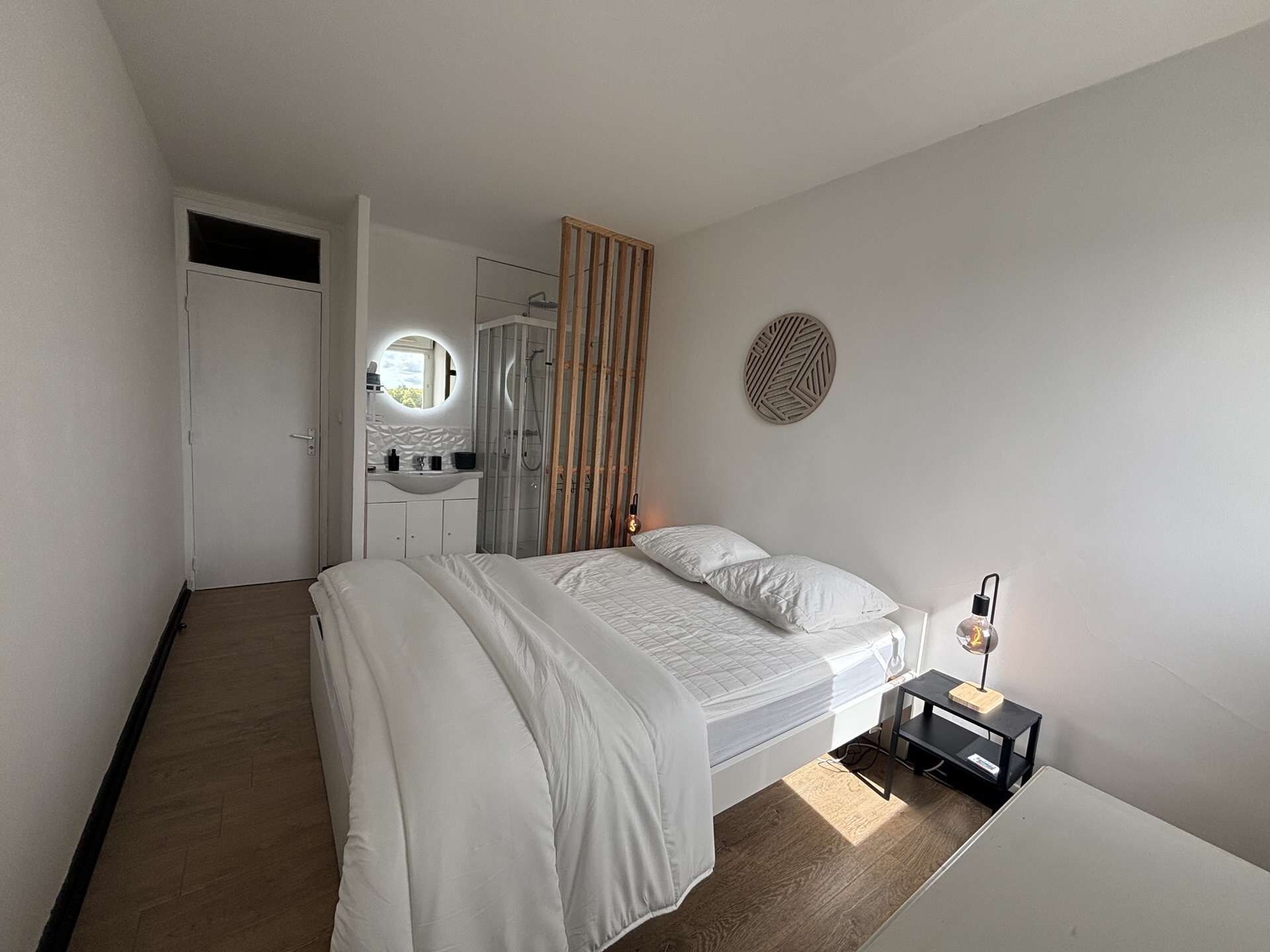 A bedroom with a bed, a bedside table, a mirror, and a shower area.