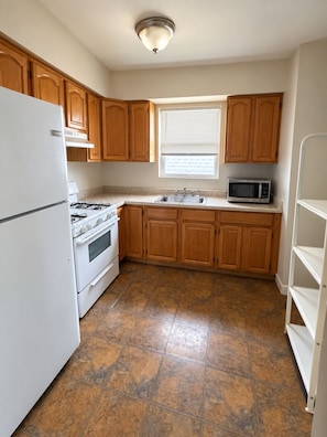 A kitchen with wooden cabinets, a white refrigerator, a stove, a microwave, and a window with blinds. - Luxury 3BR Queens Getaway; Smart TVs, A/C & Near JFK, LGA and UBS Arena (St Albans)