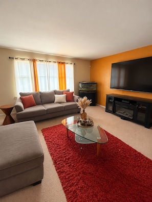 A living room with a sofa, ottoman, glass coffee table, and a red rug. - Luxury 3BR Queens Getaway; Smart TVs, A/C & Near JFK, LGA and UBS Arena (St Albans)
