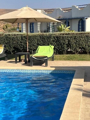 A swimming pool with lounge chairs and an umbrella, surrounded by a hedge and a building with a tiled roof.