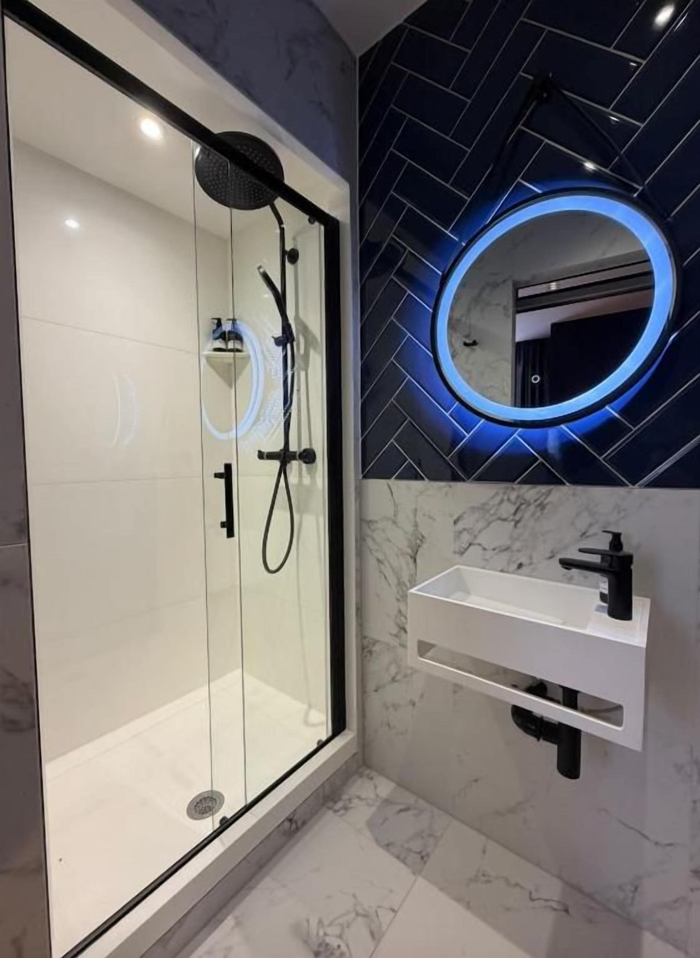 A modern bathroom with a glass-enclosed shower, a white sink with a black faucet, and a wall-mounted mirror with a blue illuminated frame.