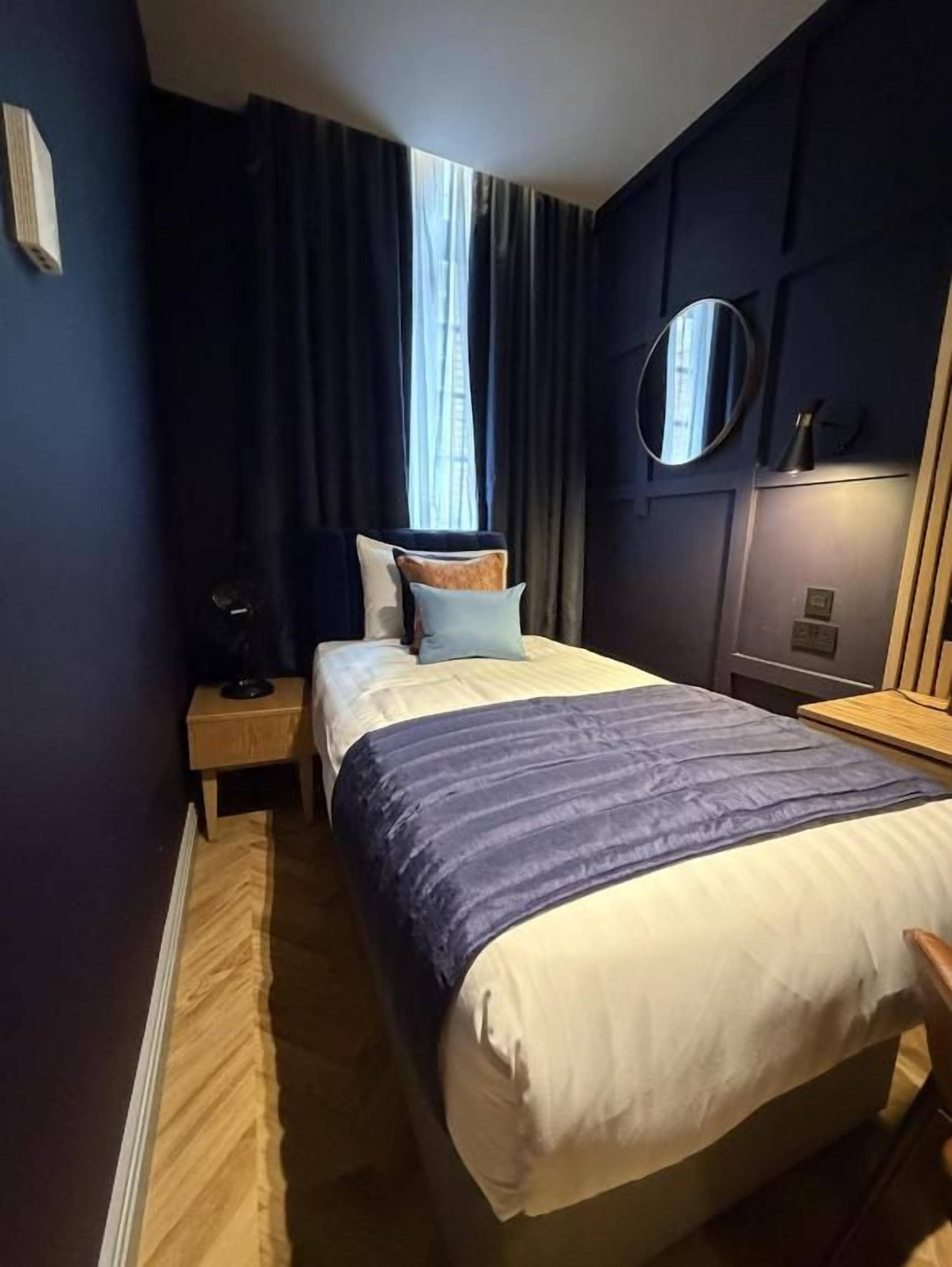 Una habitación de hotel con cama, mesita de noche, lámpara, espejo y ventana con cortinas.