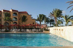 Ein Swimmingpool mit Liegestühlen und Sonnenschirmen, ein Gebäude mit Rundbogenfenstern und Palmen.