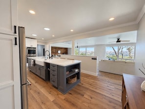 A modern kitchen with a large island, built-in appliances, and a view to a living area with a sofa and a ceiling fan.