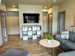 A modern living room with a grey sofa, a round coffee table with a plant, a built-in shelving unit, and a flat-screen TV mounted on the wall. - Wapato Waters-your Manson getaway-lakefront, family-friendly, dock, and hot tub. (Manson)