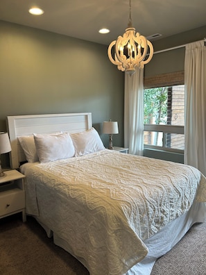 A bedroom with a large bed, a nightstand with a lamp, a chandelier, and a window with curtains. - Wapato Waters-your Manson getaway-lakefront, family-friendly, dock, and hot tub. (Manson)