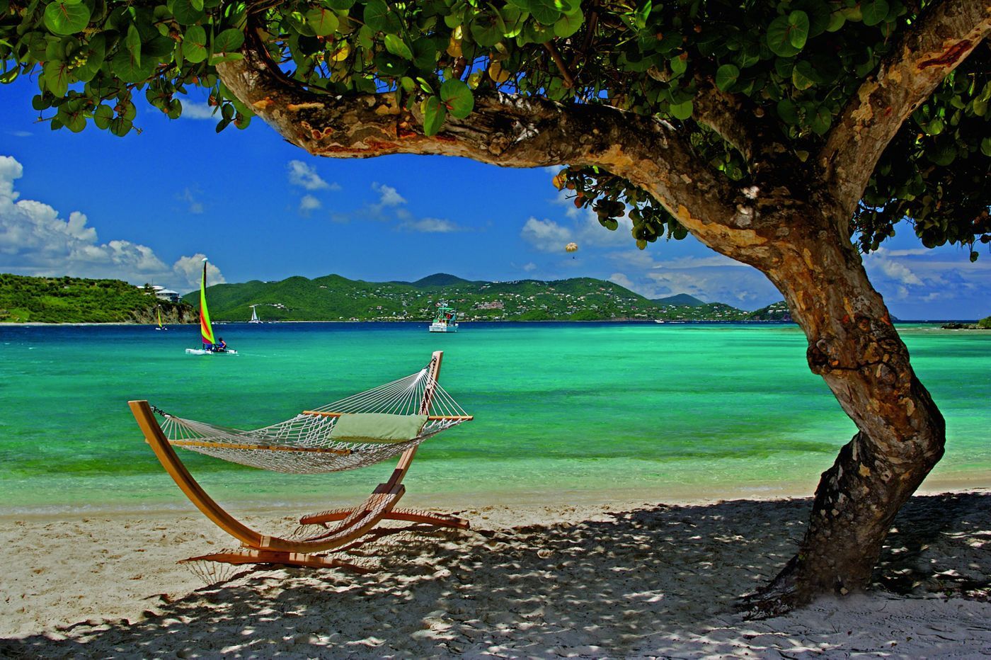 A hammock under a tree by a clear turquoise sea with boats in the distance.